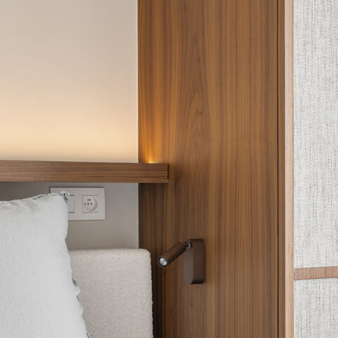Detail van een leeslampje op een notelaar paneel naast het bed, met een verlichte plank en natuurvezel kastdeur.