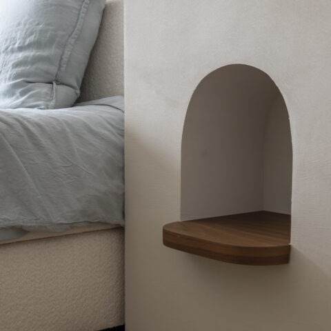 Detail van een boogvormige nachtnis in kalkpleister met een afgerond notelaar plankje naast het bed.