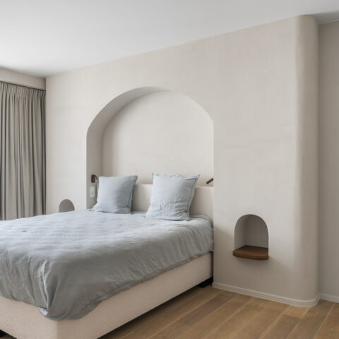 Slaapkamer met een gebogen bedwand in kalkpleister, een grote boognis rond het bed en kleine nissen met notelaar plankje.