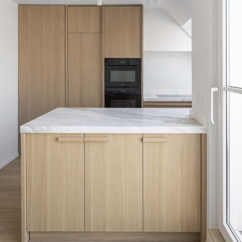 Keuken in lichte eik met hoge kasten, inbouwovens en een schiereiland met wit geaderd marmeren blad.