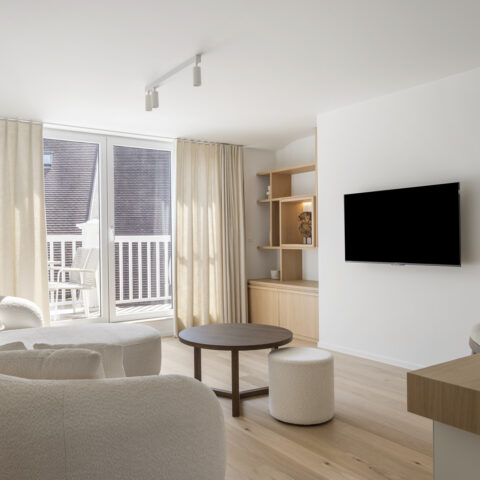 Leefruimte met een wandkast in lichte eik met open vakken naast de tv-wand en een gebogen bank bij het raam, maatwerk van YVES.