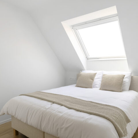 Zolderslaapkamer met hellende wand, dakraam en een gestoffeerd bed in natuurlijke tinten.