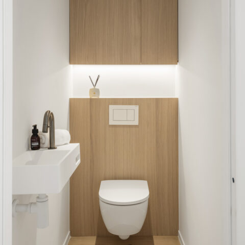 Toilet met eiken wandbekleding achter het hangtoilet, een eiken bovenkast en indirecte ledverlichting op de tablet.