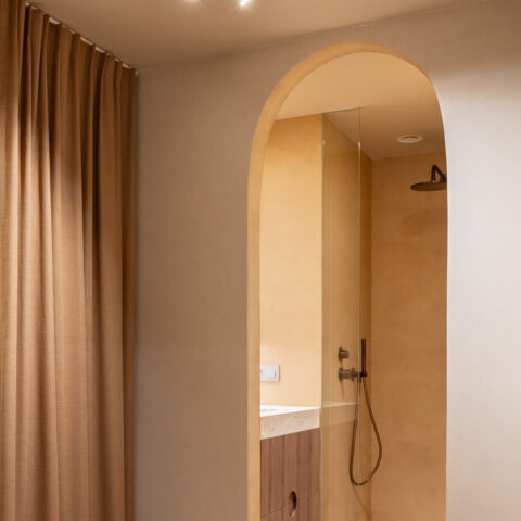 Boogvormige doorgang van de slaapkamer naar een warme badkamer met inloopdouche en notelaar badmeubel.