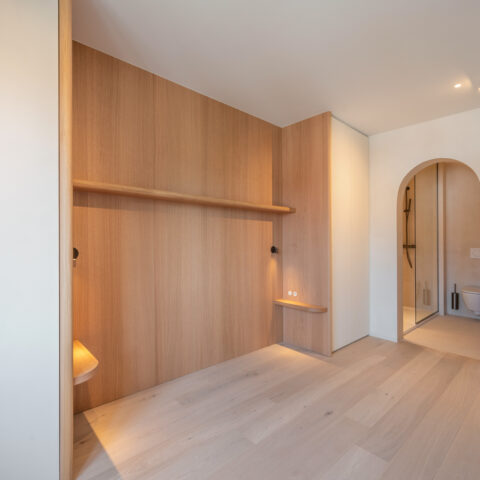Slaapkamer met eiken wandbekleding, doorlopende legplank en zwevende nachttafeltjes, met een boogdoorgang naar de badkamer, maatwerk van YVES Interieur Op Maat.