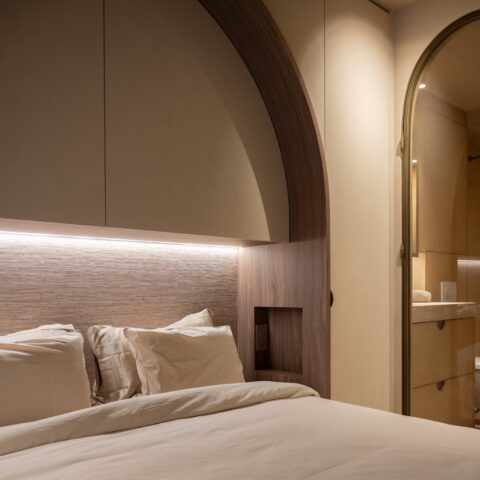 Bed onder een boogvormige kastwand met ledstrip, nachtnis in notelaar en zicht op de badkamer via een gebogen glazen deur.