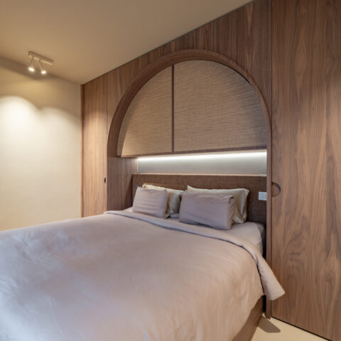 Slaapkamer met kastwand in notelaar rond een boogvormige nis met natuurvezelpanelen en ledverlichting boven het bed.