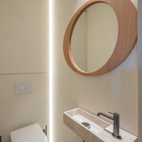 Toilet met een travertin fonteintje, ovale spiegel in eik en een verticale lichtlijn in de wand.